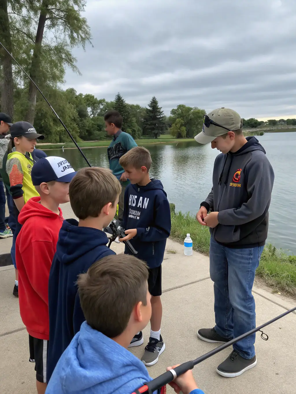 A group of children laughing and learning how to cast fishing lines at the edge of a pond during an O'FIL DE L'O fishing day event.